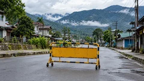 A quiet generic eastern Himalayan town street with lush foothills, a simple unattended portable road barrier in foreground, overcast daylight. No fires, riots, police confrontation or identifiable incident; conceptual public order illustration.